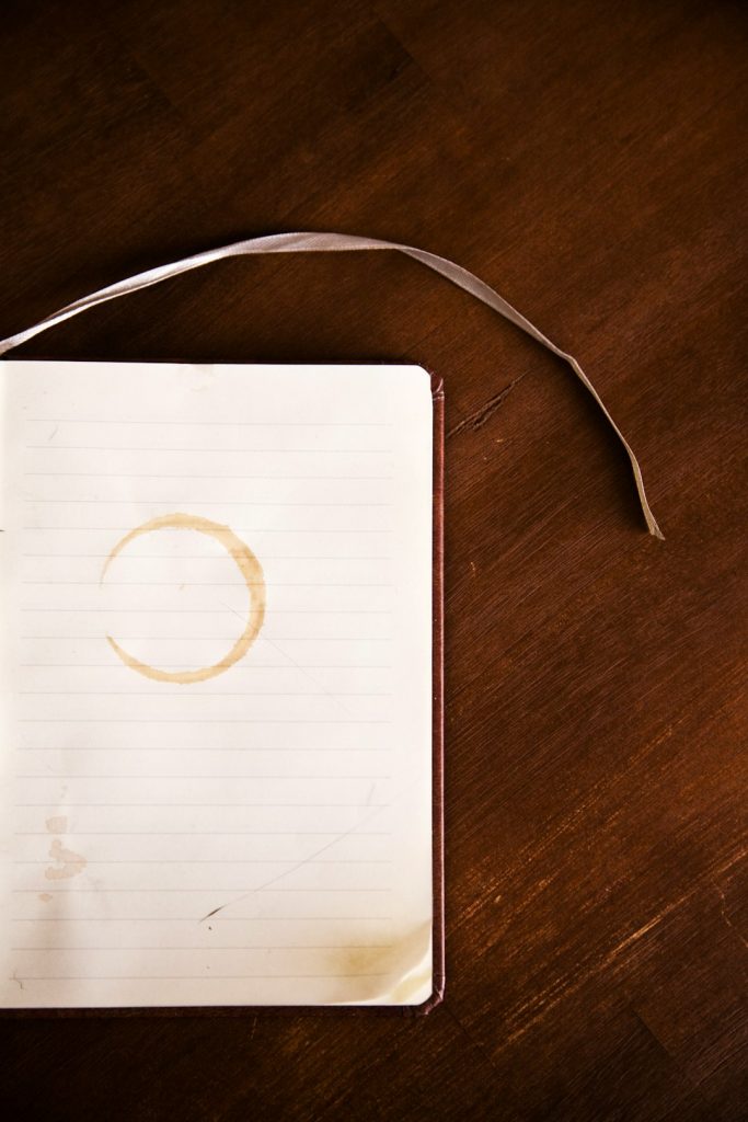 Open notebook on a dark wooden table with a coffee ring stain on the lined page and a white ribbon bookmark.