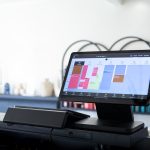 Touchscreen tablet on a cash register displaying a colorful weekly calendar/schedule app in a bright store environment