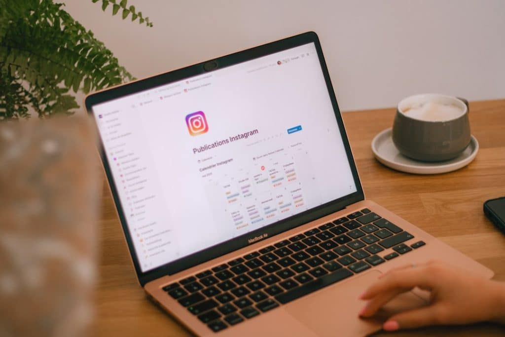 MacBook Air on a wooden table shows an Instagram page, with a cup of coffee on a saucer to the right and a plant in the background.