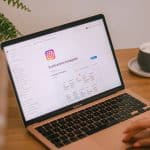 MacBook Air on a wooden table shows an Instagram page, with a cup of coffee on a saucer to the right and a plant in the background.