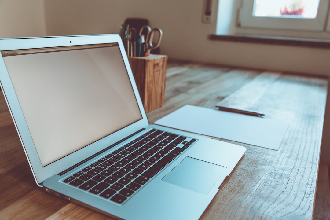 Open silver laptop on a wooden desk with a blank sheet of paper and a pencil nearby in a bright room by a window.