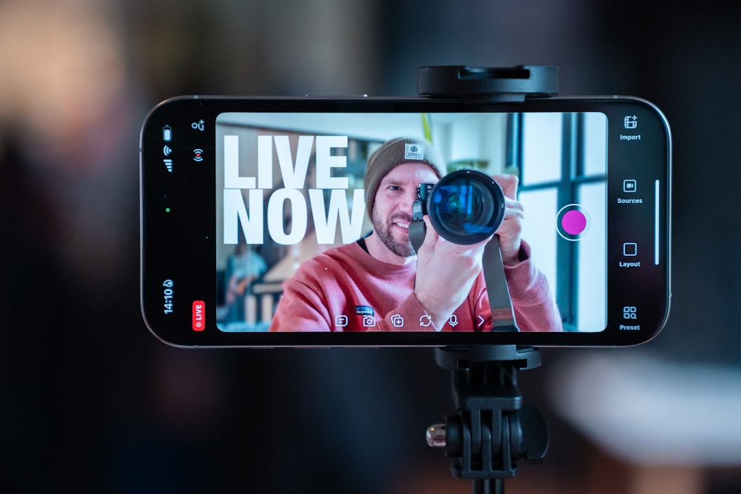 Smartphone on a tripod showing a man with a camera lens, with the words 'LIVE NOW' on the screen.