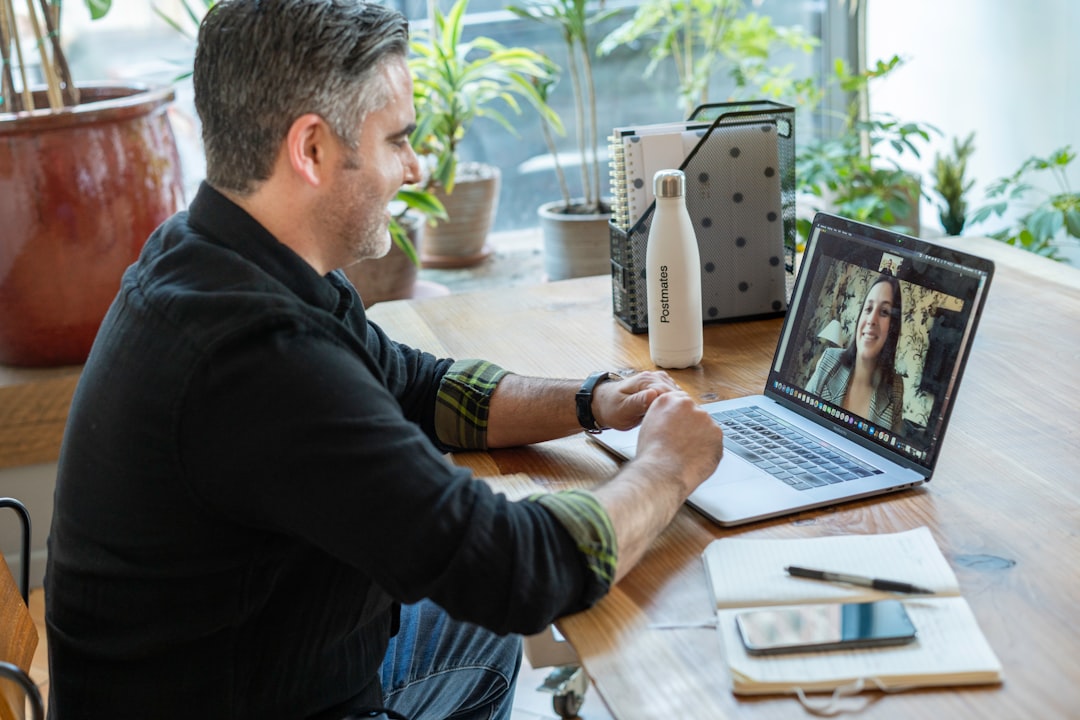 Man in a dark shirt sits at a wooden desk, engaged in a video call on a laptop showing a smiling woman on screen with plants in the background on a bright home office.