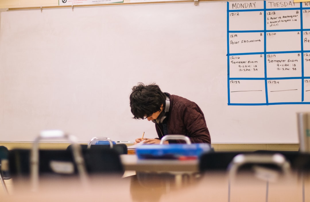 Student wearing headphones sits at a desk in a classroom, writing with a pencil, with a blue weekly calendar on the whiteboard behind him.