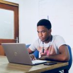 Young man in a gray t‑shirt sits at a desk typing on a laptop, headphones around his neck, in a classroom setting.