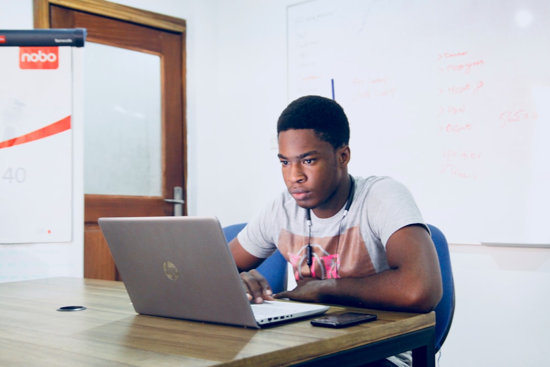 Young man in a gray t‑shirt sits at a desk typing on a laptop, headphones around his neck, in a classroom setting.