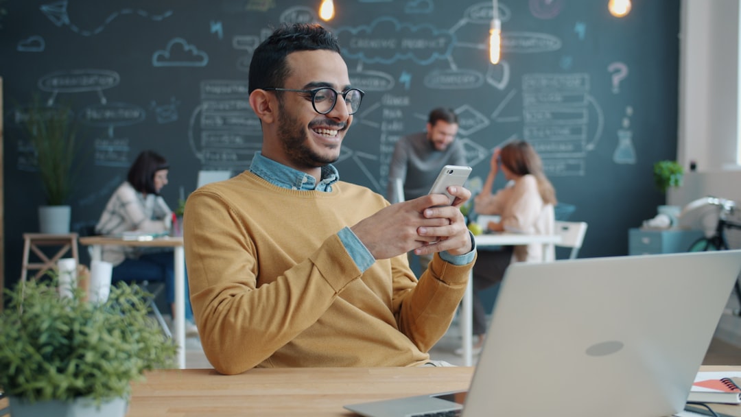 Young man with glasses in a mustard sweater sits at a table in a coworking space, smiling as he uses his phone.