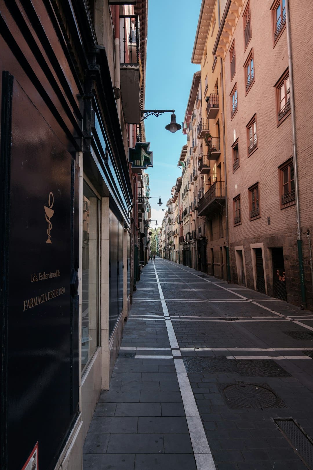 Narrow European street with tall pastel buildings, a pharmacy on the left, and a blue sky above.”