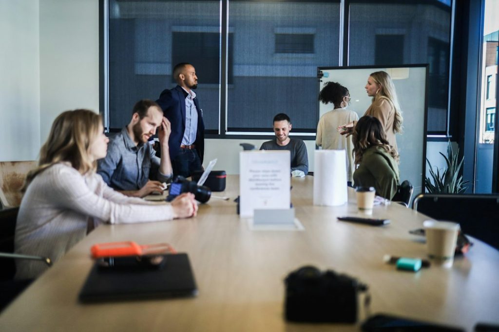 Group of colleagues around a conference table in a bright office, some with laptops and notebooks, others talking, with a whiteboard/tv screen in the background.