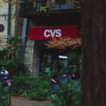 CVS storefront with a red sign, surrounded by plants and a few people wearing masks nearby.
