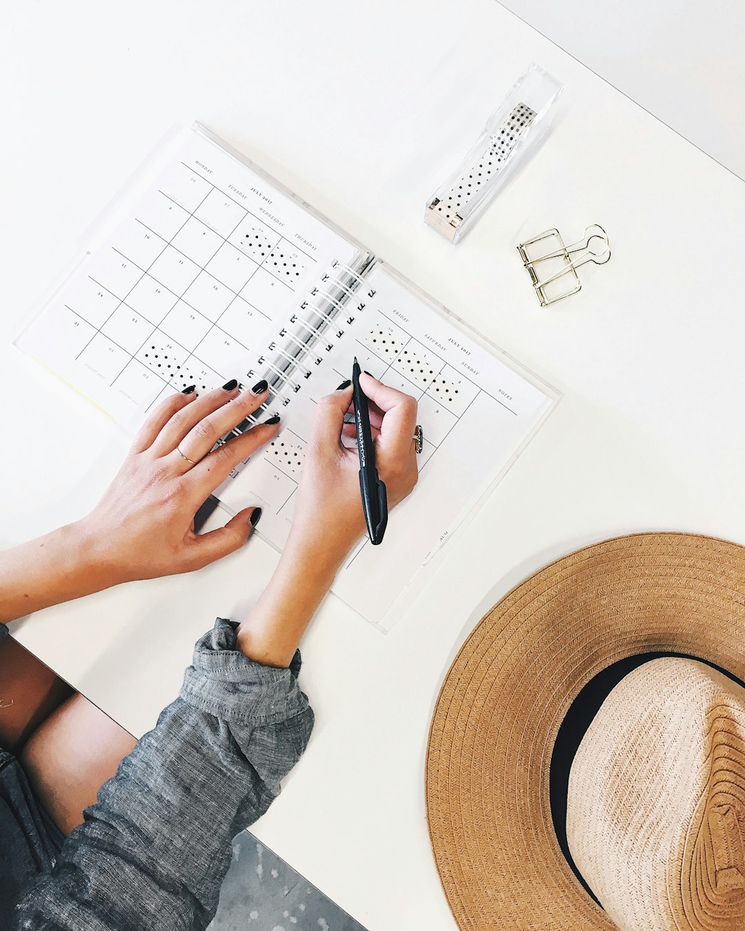 Person writing in a spiral-bound planner on a white desk; a wide-brim straw hat sits nearby.