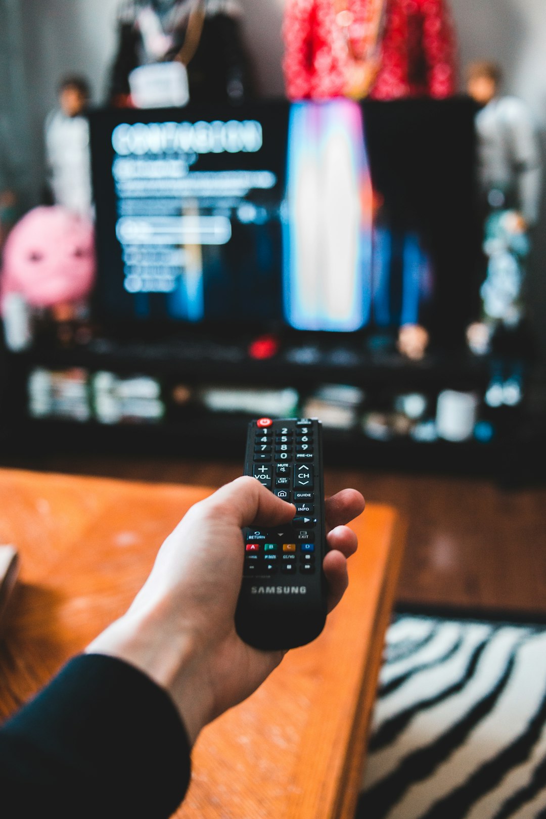 Hand holding a Samsung TV remote aimed at a blurred TV screen showing a connection/setup menu in the background.