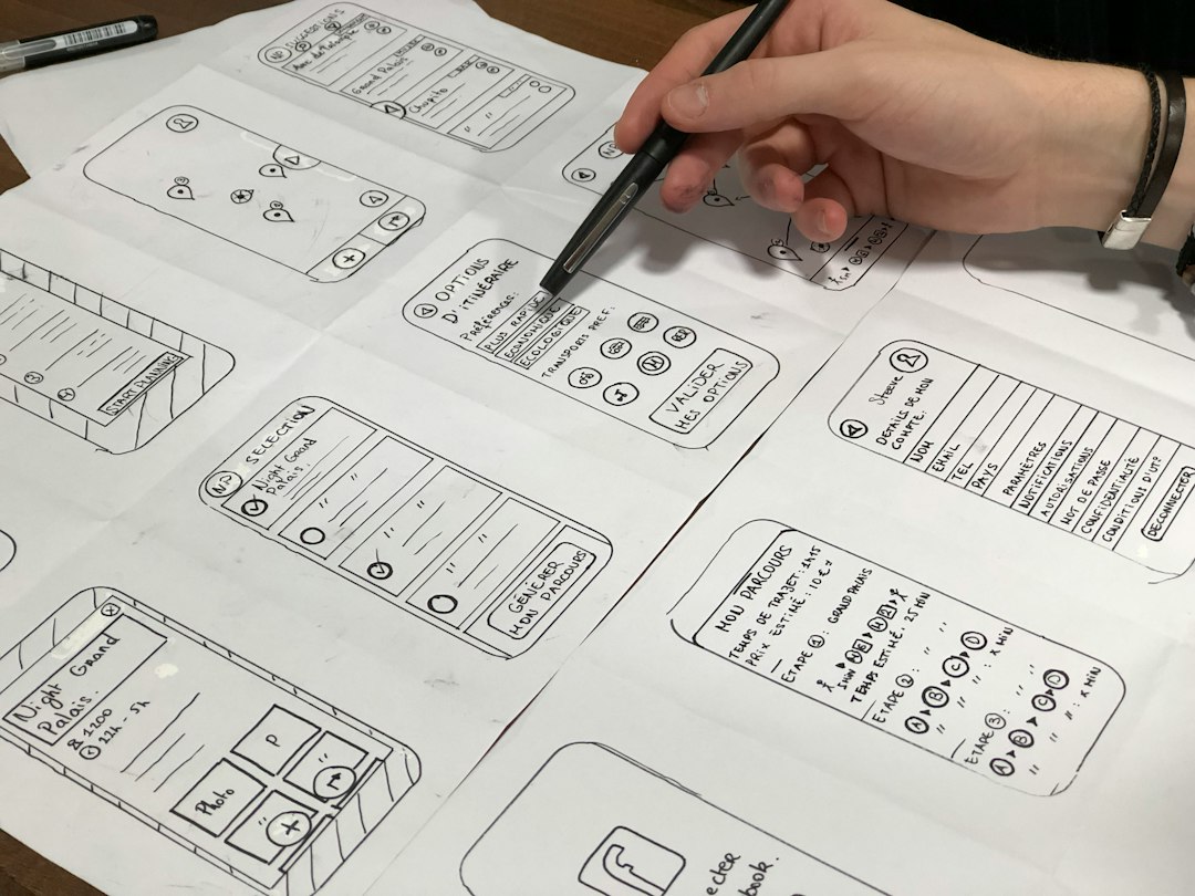 A person sketches mobile app wireframes on scattered sheets of paper, using a pen to draw button layouts and UI elements across several designs.