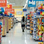 Person walks down a brightly lit supermarket aisle with promotional signs and tall shelves on both sides.