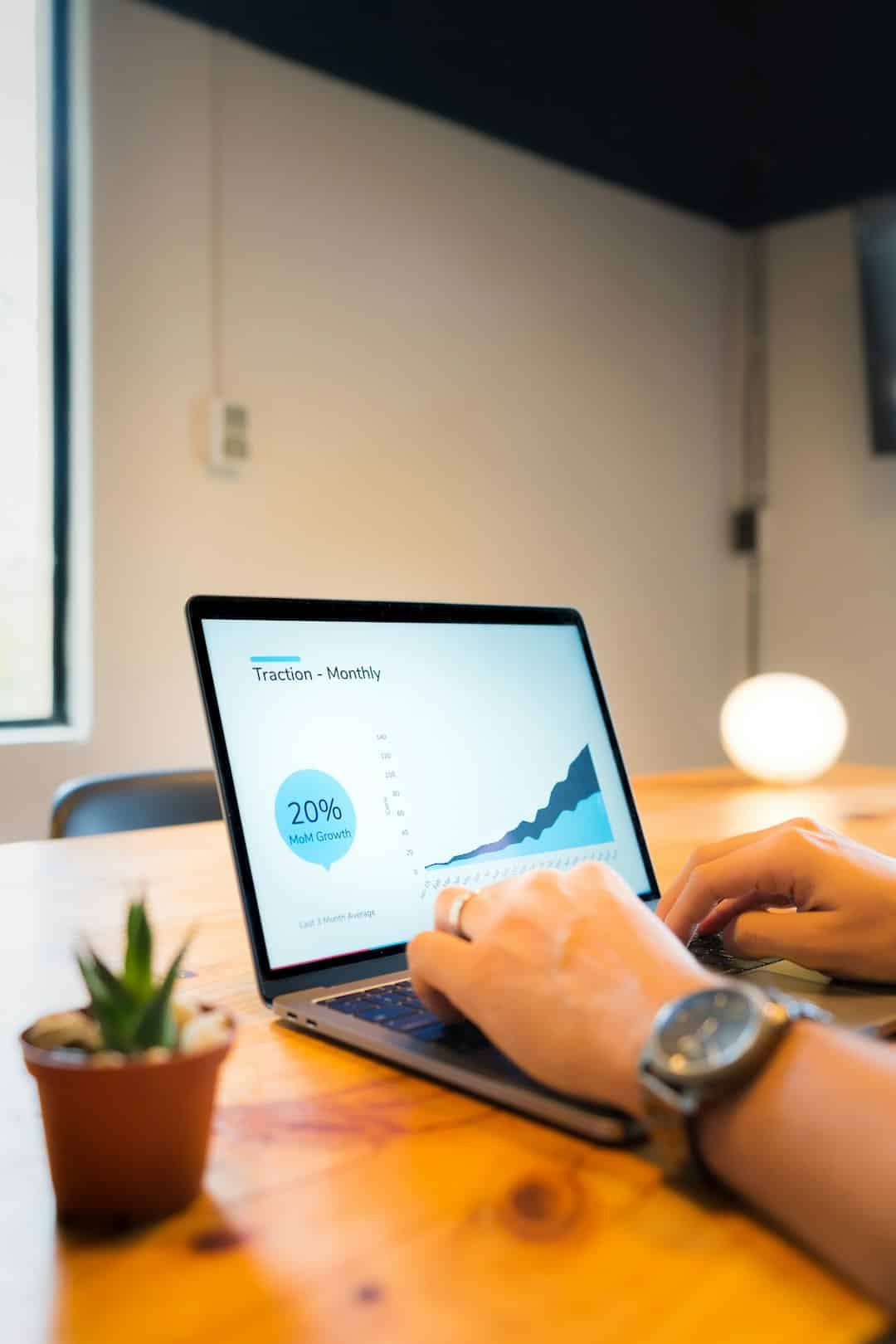 Person typing on a laptop at a wooden table with charts on the screen and a small potted plant nearby.