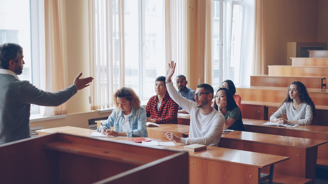 Professor lecturing at the front as students in a classroom listen; one student raises a hand to ask a question.