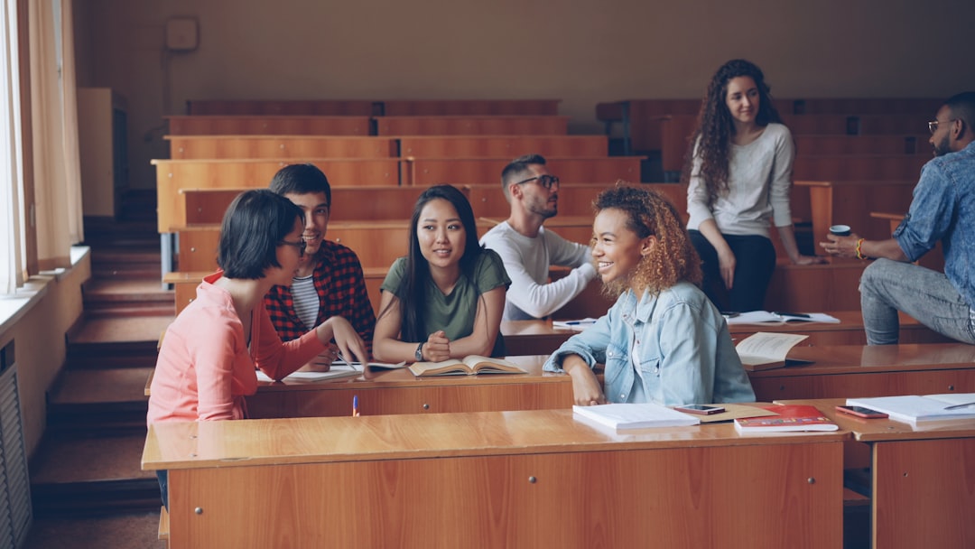 Group of diverse students chatting at wooden desks in a university lecture hall, with notebooks and books open around them.