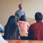 Teacher stands at front of classroom delivering lesson to seated students.