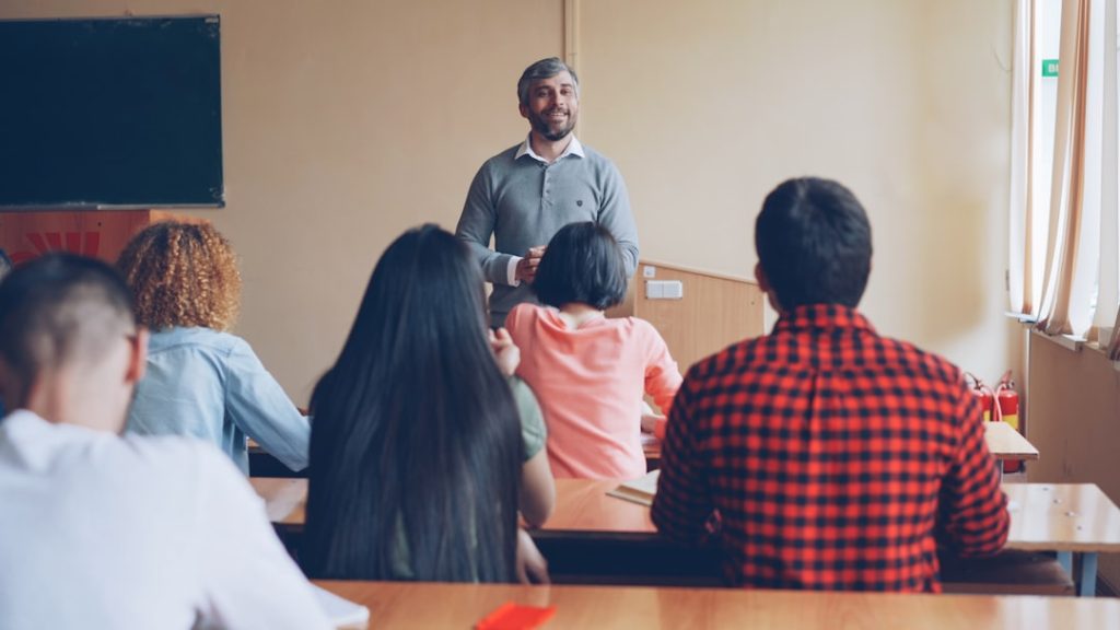 Male teacher standing at the front of a classroom addressing students seated at desks, smiling.