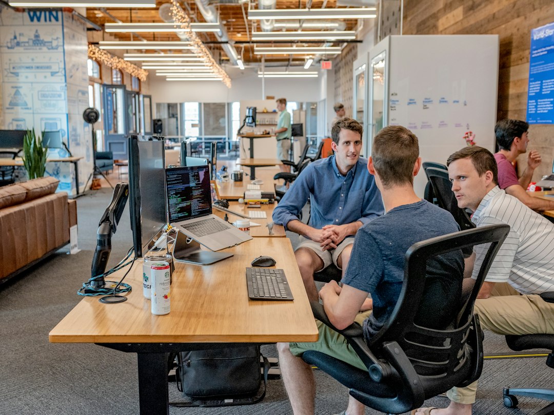Three coworkers at a long shared desk in a bright open office, laptops and multiple monitors as they chat nearby a beverage can on the desk.