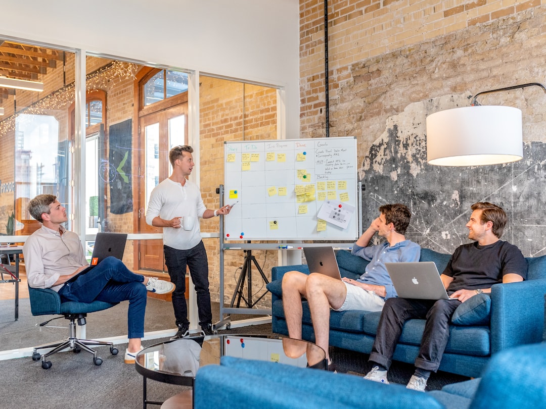 A man stands at a whiteboard with sticky notes while three colleagues listen in a casual office lounge.