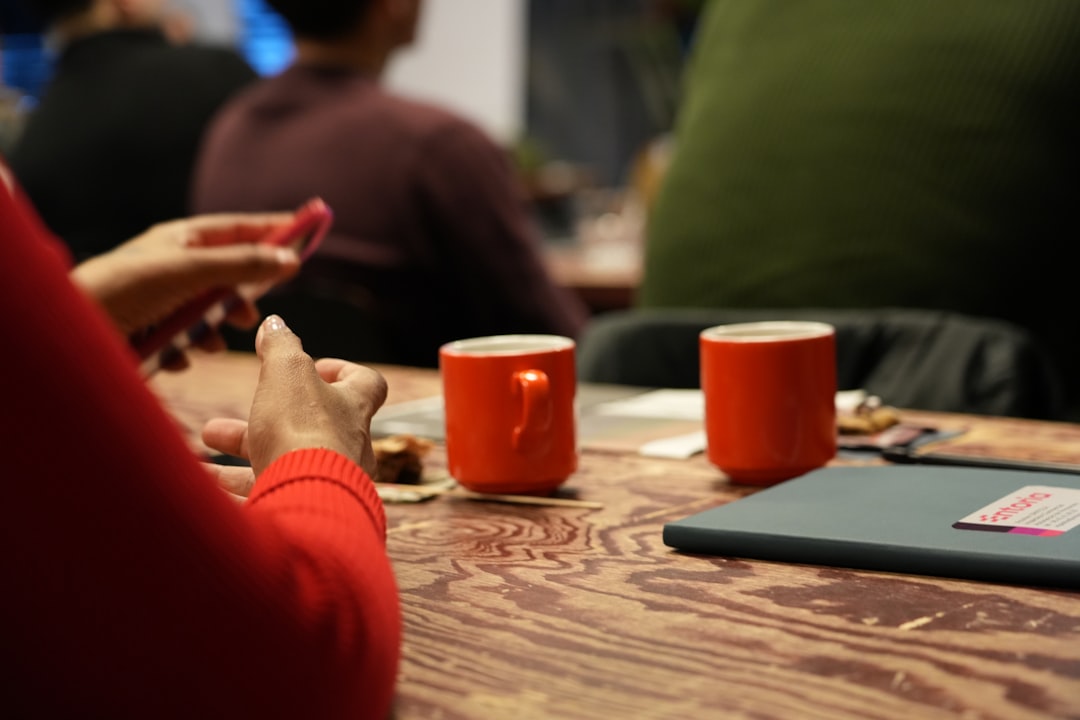 Two orange mugs sit on a patterned wooden table in a busy cafe, with a person in a red sweater using a phone in the foreground.