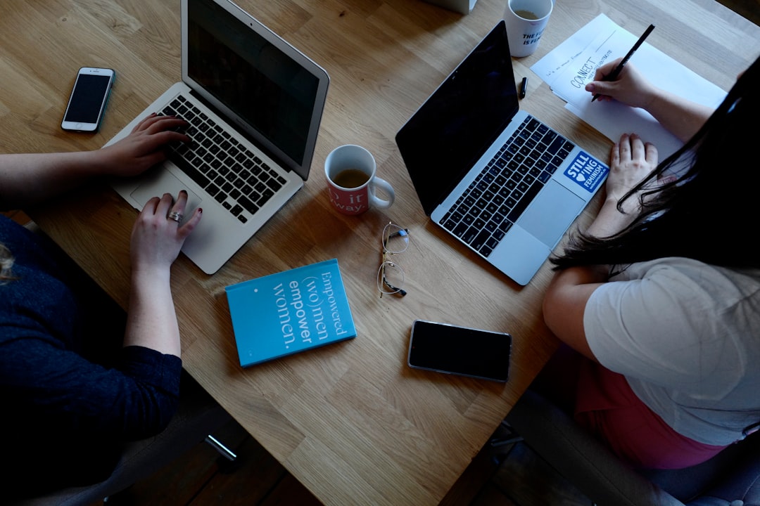 Overhead view of two people working at a wooden table with laptops, a blue book, coffee mugs, glasses, and phones on the desk.