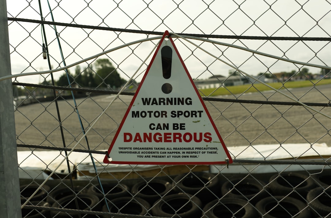 Triangular warning sign on a chain-link fence at a racetrack: 'WARNING MOTOR SPORT CAN BE DANGEROUS' with additional small text below.