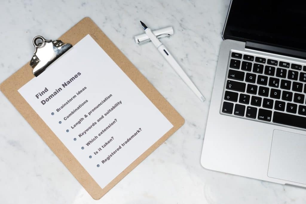 Clipboard note titled 'Find Domain Names' with brainstorming bullets beside a laptop on a marble desk.