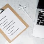 Clipboard note titled 'Find Domain Names' with brainstorming bullets beside a laptop on a marble desk.