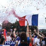 Crowd of cheering people at a celebration, waving a large French flag as pink smoke and colorful confetti fill the air.