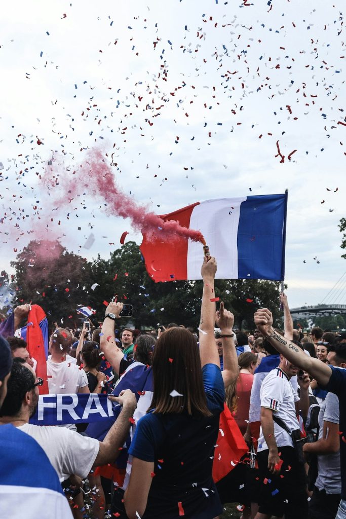woman holding red smoke can cyber shield france flag devices - AI Bud Crowd of cheering people at a celebration, waving a large French flag as pink smoke and colorful confetti fill the air.