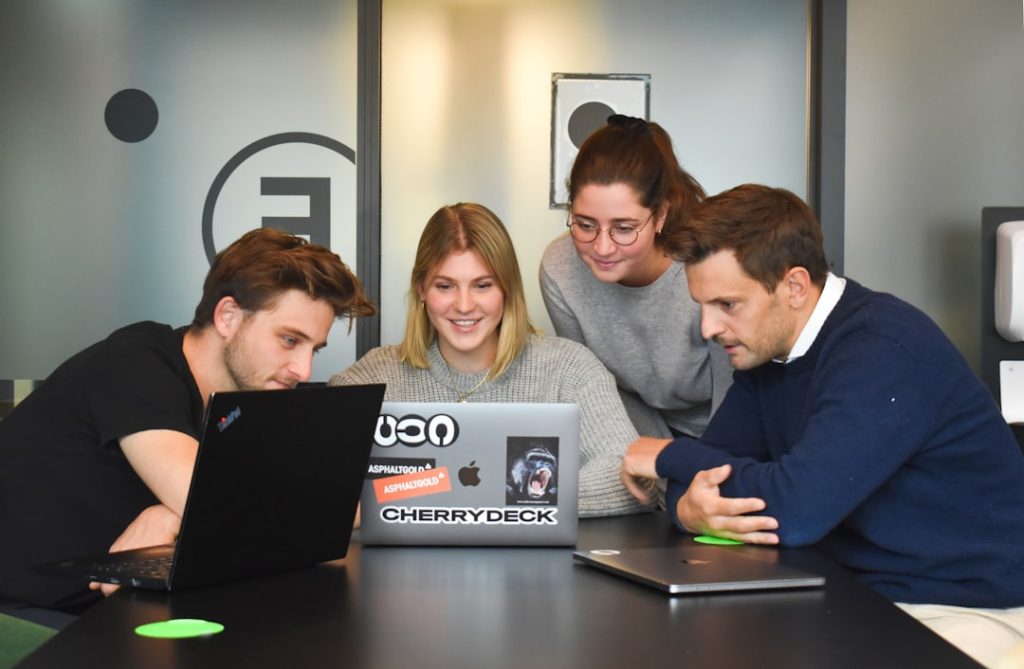 Four coworkers gathered around a table, looking at laptops and discussing a project in a modern office setting.