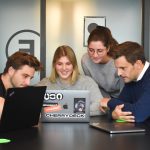 Four coworkers gathered around a table, looking at laptops and discussing a project in a modern office setting.