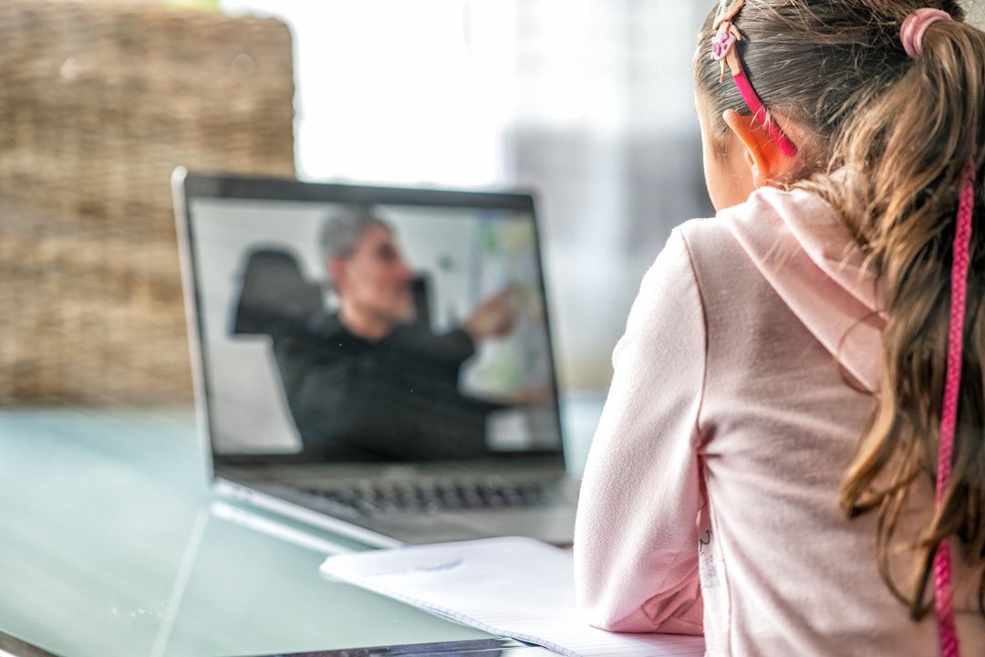 Child in a pink hoodie sits at a notebook, watching a video call on a laptop with a man gesturing on screen