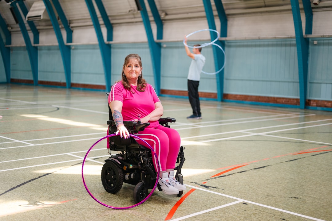 Woman in a pink tracksuit sits in a wheelchair in a gym, while a man in the background spins a hula hoop.