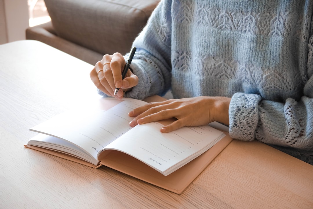 Person writing in an open notebook on a wooden desk, wearing a light blue knitted sweater.