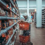 Elderly woman wearing a beret and sweater reads a bag of bread while pushing an orange shopping cart down a supermarket aisle.