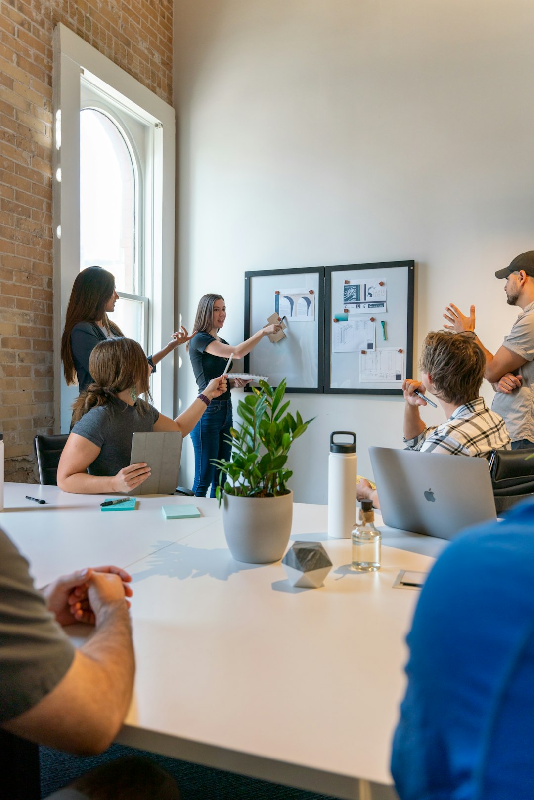 Five coworkers in a bright loft-style office present plans on two framed boards while others listen around a white conference table with laptops and notes.