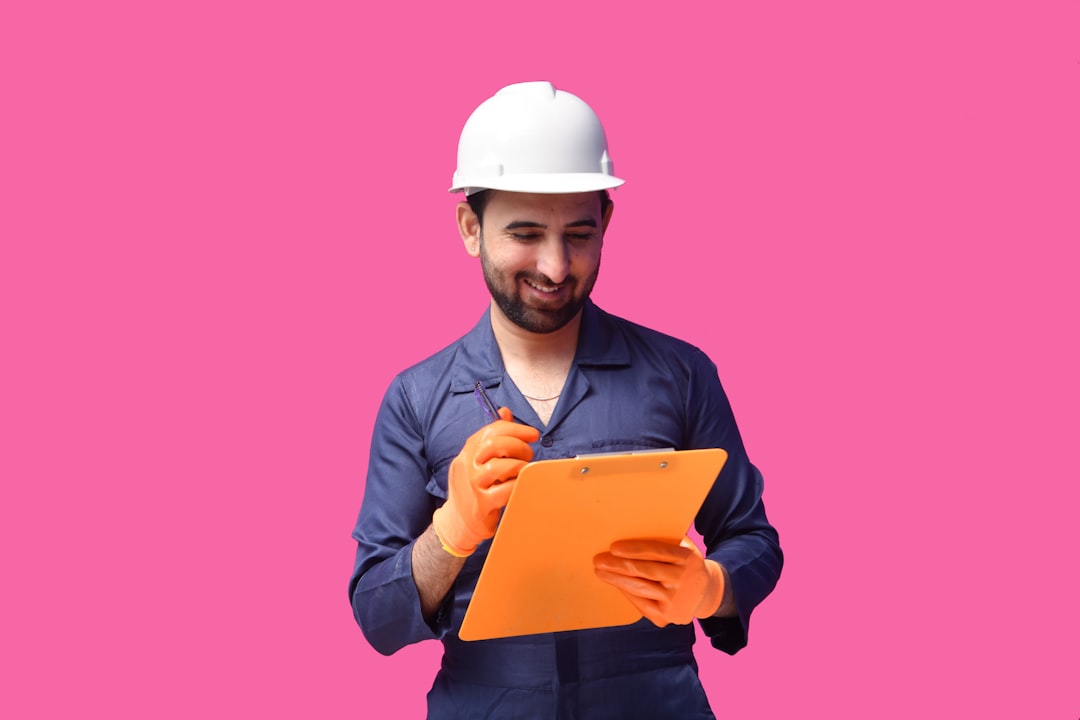 Construction worker in a white hard hat and navy coveralls writes on an orange clipboard.