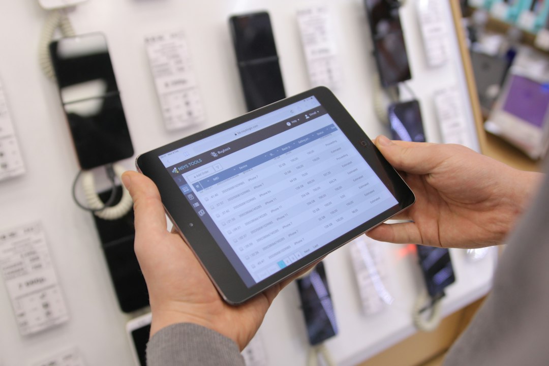 Person holds a tablet in landscape mode displaying a grid of data in a tech store setup with smartphones on the wall behind.