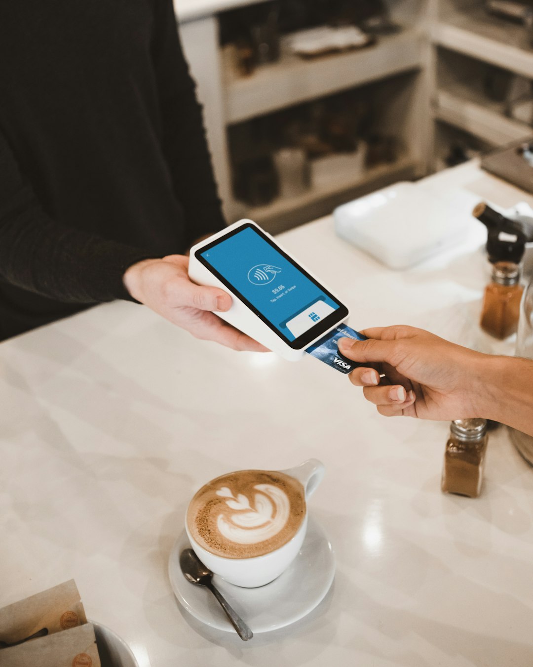 Person uses a contactless payment device to tap a card at a cafe counter, with a latte on the table nearby.