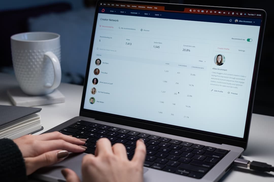 Person typing on a laptop displaying an analytics dashboard with metrics, while a white mug and stacked notebooks sit nearby on the desk