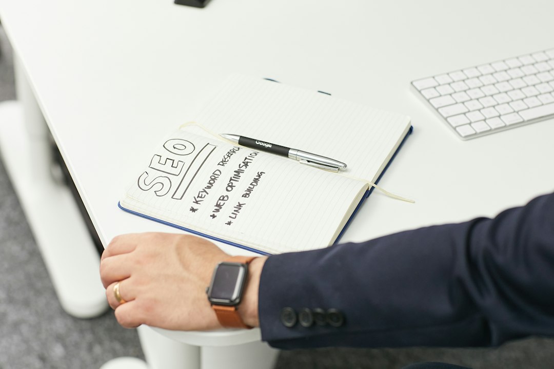 Open notebook on a white desk with a pen and SEO notes, a smartwatch on a person’s wrist, and a keyboard in the background.