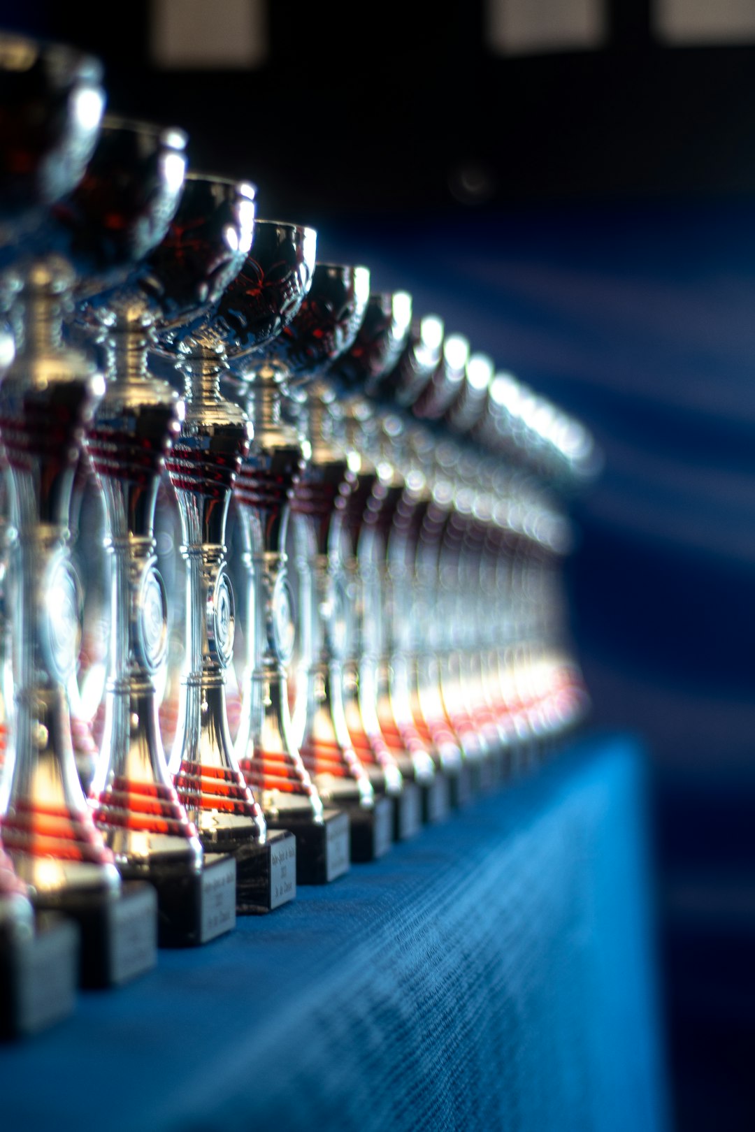 Row of shiny trophies lined up on a blue table, extending into the distance, with a shallow depth of field showing highlights on the cups
