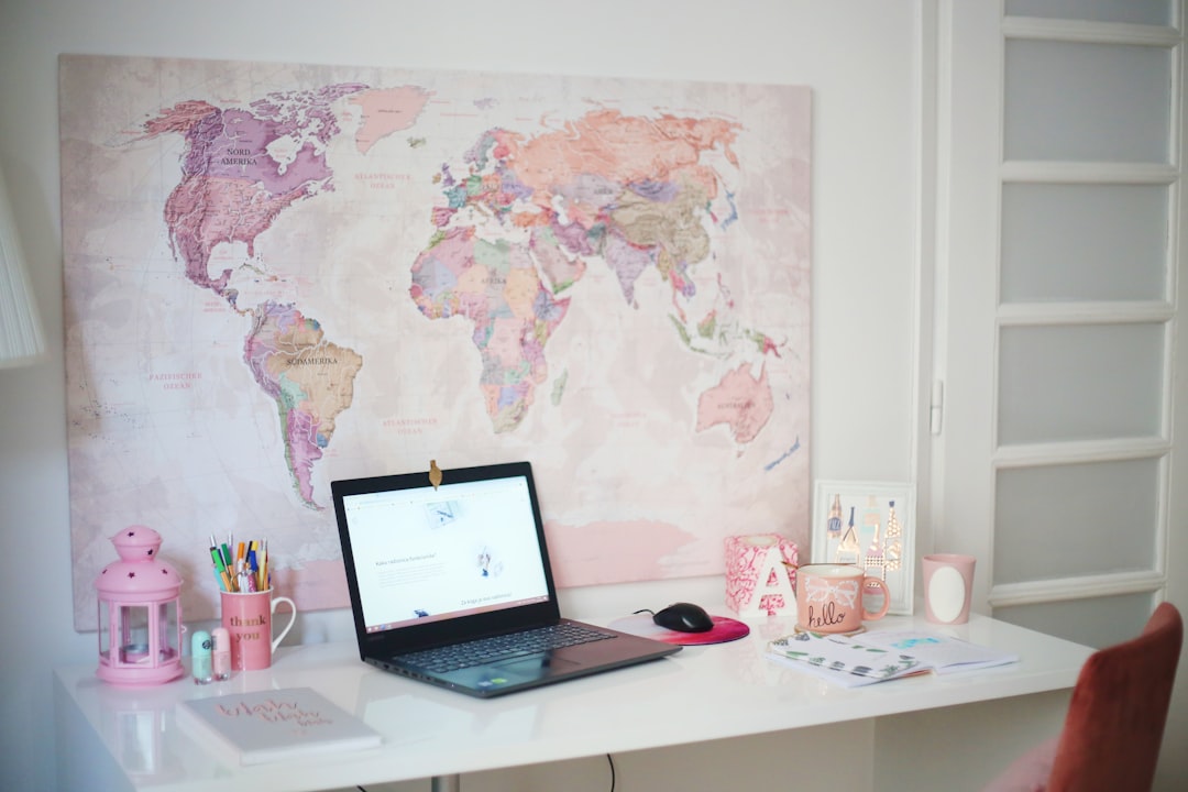 Desk setup with a pastel pink theme, laptop open on a white desk in front of a large world map wall art.