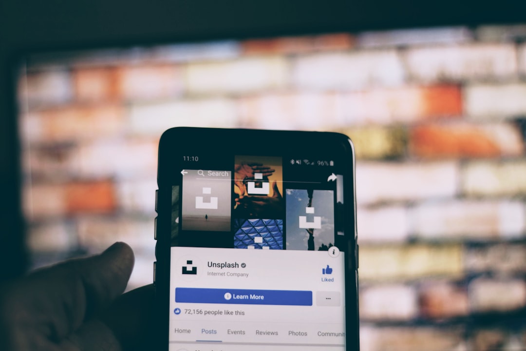 Close-up of a hand holding a smartphone showing the Unsplash Facebook page on the screen, with a blurred brick wall in the background.