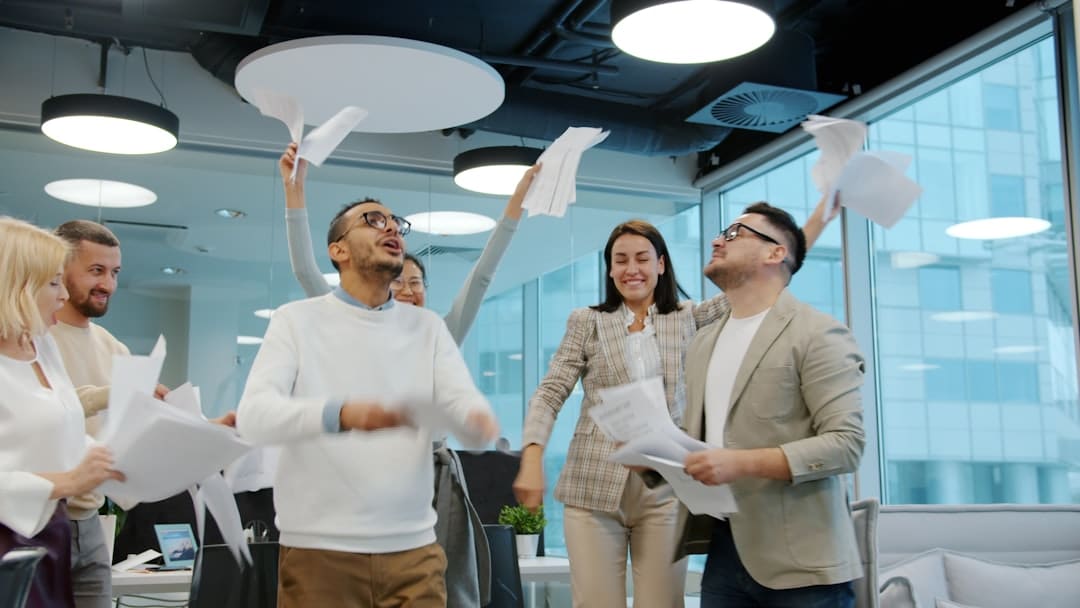 Diverse coworkers celebrate in a modern office, throwing papers in the air and smiling near glass walls and round ceiling lights.