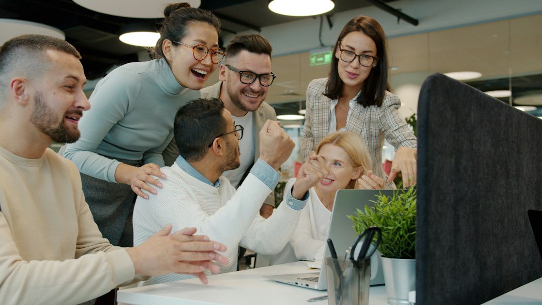 Diverse team of six coworkers gathered around a laptop, celebrating with smiles in a bright office setting.