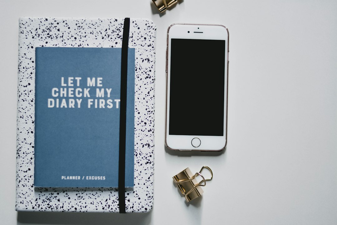 Blue planner with the words 'LET ME CHECK MY DIARY FIRST' sits on a speckled notebook, beside a smartphone and gold binder clips on a light desk surface.